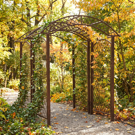 H Potter brand dark brown metal arched garden trellis with lattice panels, shown in a lush autumn garden with yellow and orange foliage.