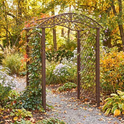 A dark brown H Potter metal garden arch with lattice panels, covered in climbing green vines and small purple flowers, situated on a gravel path in a lush autumn garden.