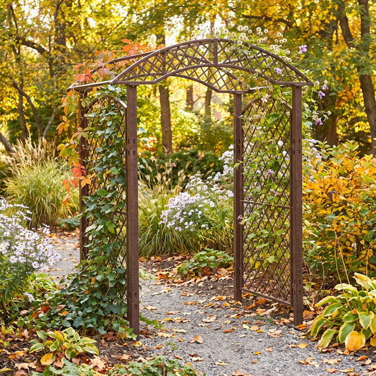 A dark brown H Potter metal garden arch with lattice panels, covered in climbing green vines and small purple flowers, situated on a gravel path in a lush autumn garden.