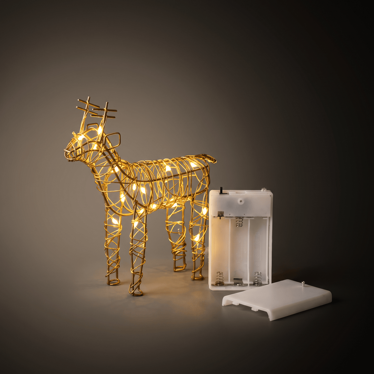 Small gold wire-frame reindeer Christmas light decoration with warm glowing bulbs, shown next to its white battery compartment and battery cover on a dark gray gradient background.
