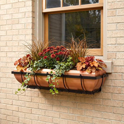 h potter brand copper rectangular window box planter with a black metal frame, filled with autumnal greenery, red flowers, and trailing ivy, mounted on a beige brick wall next to a window.