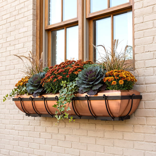 H Potter brand copper window box planter with black metal brackets, filled with orange and yellow flowers, purple cabbage, and green ivy, mounted on a beige brick wall under a wooden window.