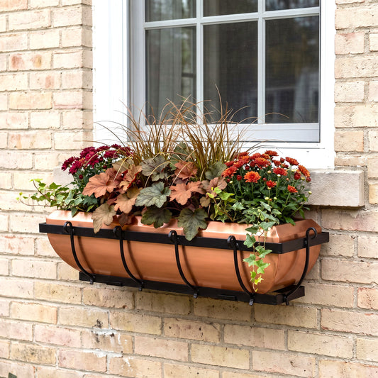 h potter copper planter box with a black metal bracket, filled with autumnal green and red ornamental plants, mounted on a light beige brick wall next to a white-framed window.