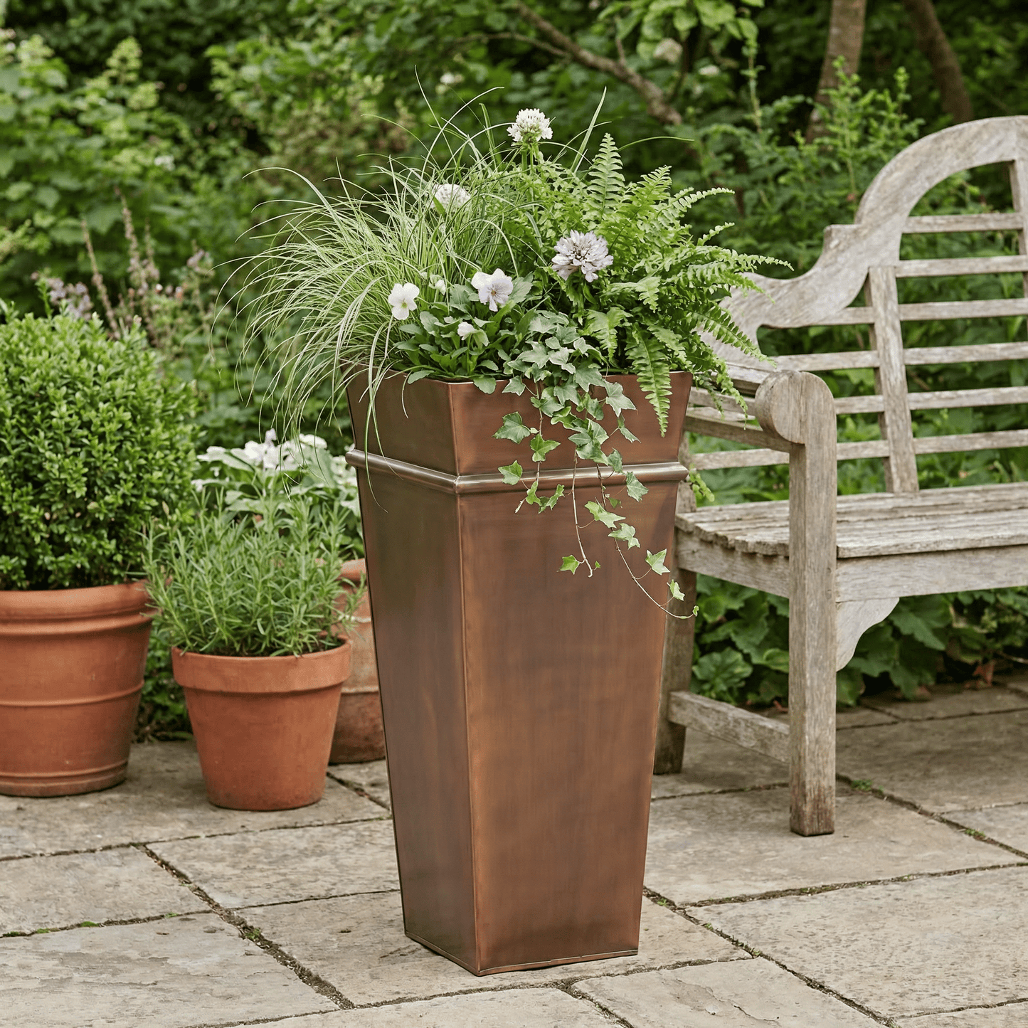 h potter tall antique copper planter on stone patio with flowers and ferns