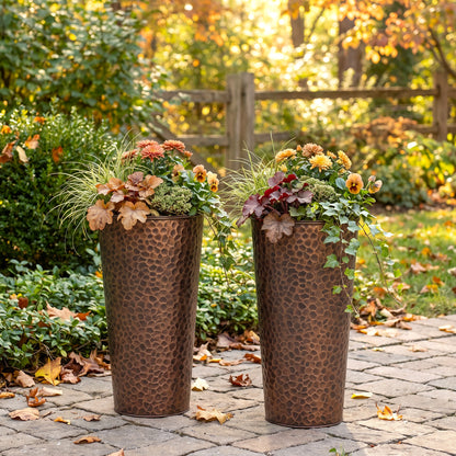 h potter brand two tall hammered metal planters in a coppery bronze finish, filled with autumnal flowers and greenery, placed on a stone pathway in a garden setting.