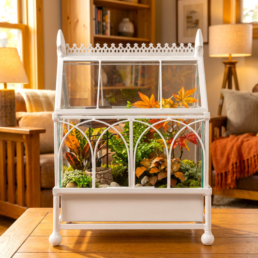 h potter brand white metal and glass terrarium display case with a decorative top and front arches, filled with small green plants and orange autumn leaves, set on a wooden table in a warm, cozy living room.