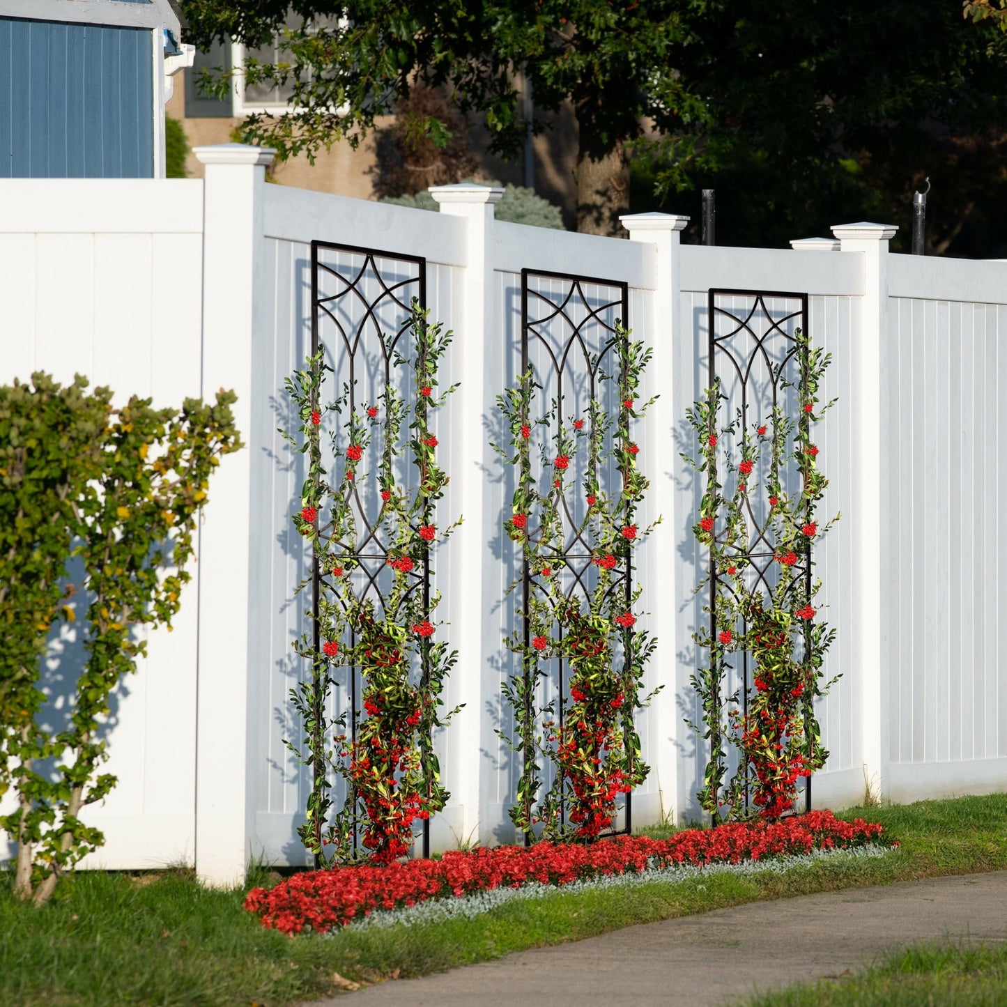 Three tall black metal garden trellises with a geometric design, set against a white fence with climbing plants.
