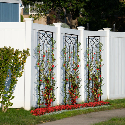 Three tall black metal garden trellises with a geometric design, set against a white fence with climbing plants.