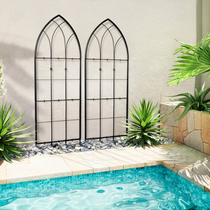 Two black iron trellises with a gothic style design, placed beside a pool in an outdoor setting with plants in the background.