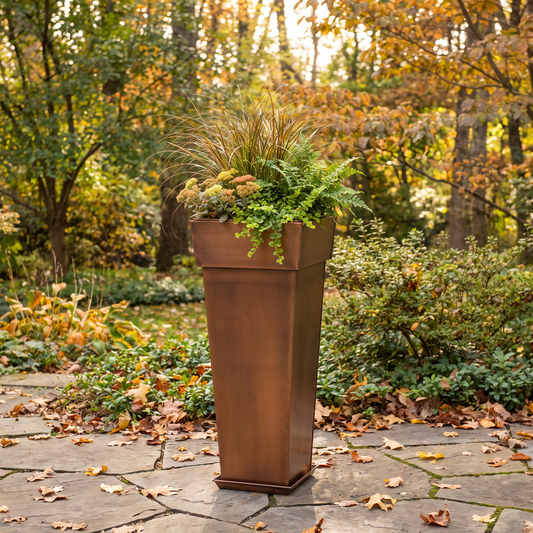 h potter brand tall rectangular copper-toned planter filled with ornamental grass and green foliage, placed on a stone patio in an outdoor garden with autumn trees in the background.