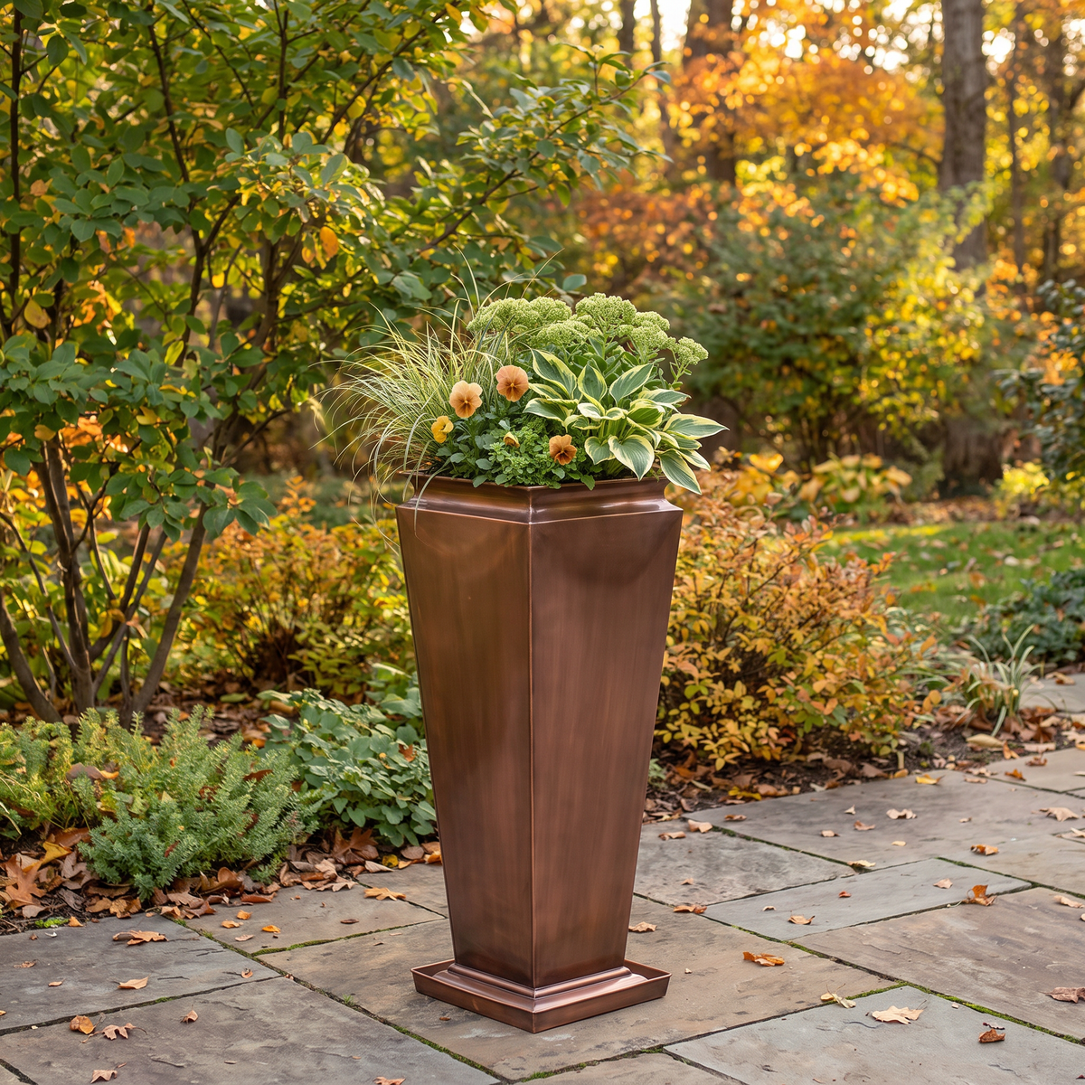 h potter brand tall planter with a tiered base, filled with green foliage and small orange flowers, set on a stone patio in an autumn garden.