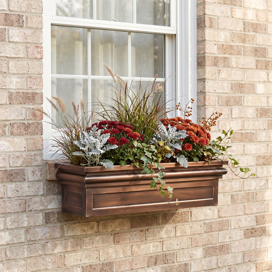 A rectangular copper-toned metal window box planter filled with autumnal flowers and ornamental grasses, mounted on a beige brick wall beneath a white-framed window.