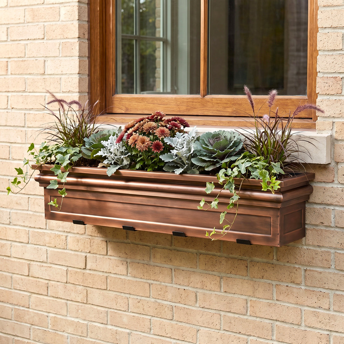 A copper-colored metal window box planter mounted on a beige brick wall beneath a wooden window, filled with green succulents, trailing ivy, and ornamental flowers.