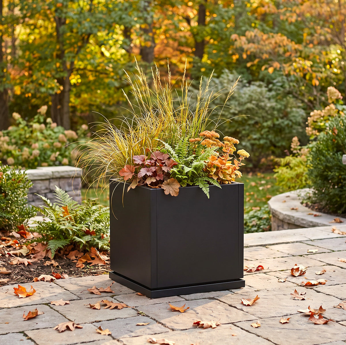 large matte black square planter box filled with ornamental grasses and autumn foliage, placed on a stone patio in an outdoor garden setting.