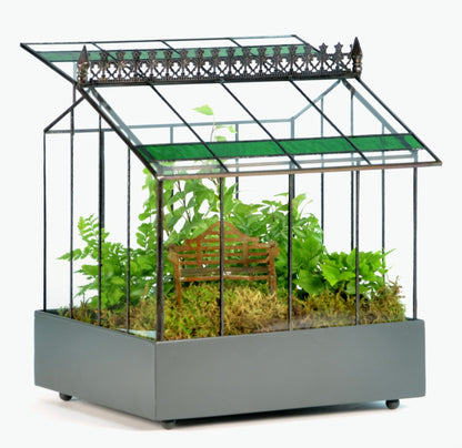 A classic H Potter glass terrarium Wardian case with a black powder-coated metal tray base and a hinged roof, planted with lush green ferns and a miniature park bench